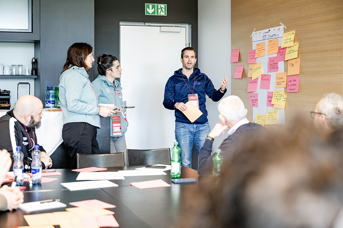 Ein Referent steht vor einer Stellwand mit vielen Post-it-Zetteln und erklärt den Zuhörenden etwas.